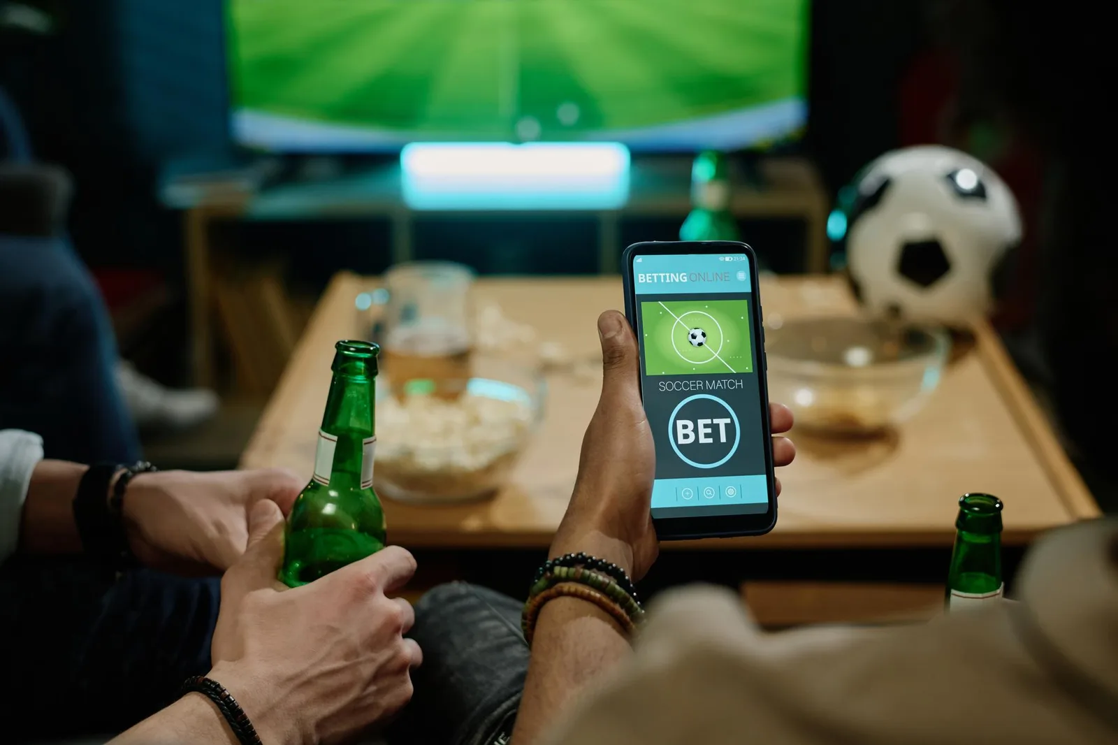 Pessoa segurando celular com aplicativo de apostas esportivas e garrafa de cerveja, enquanto assiste a jogo de futebol na TV com amigos.