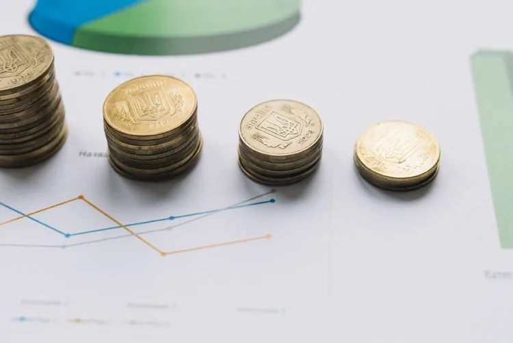 Pilhas de moedas douradas decrescentes sobre gráficos financeiros, ilustrando projeções econômicas e a reforma tributária em 2027 focada na cobrança por consumo.