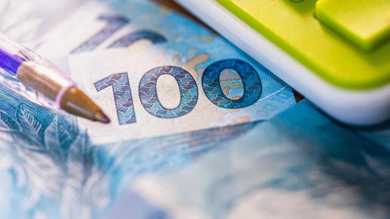Notas de cem reais, caneta e calculadora sobre a mesa, ilustrando o impacto da reforma tributária no preço dos produtos.