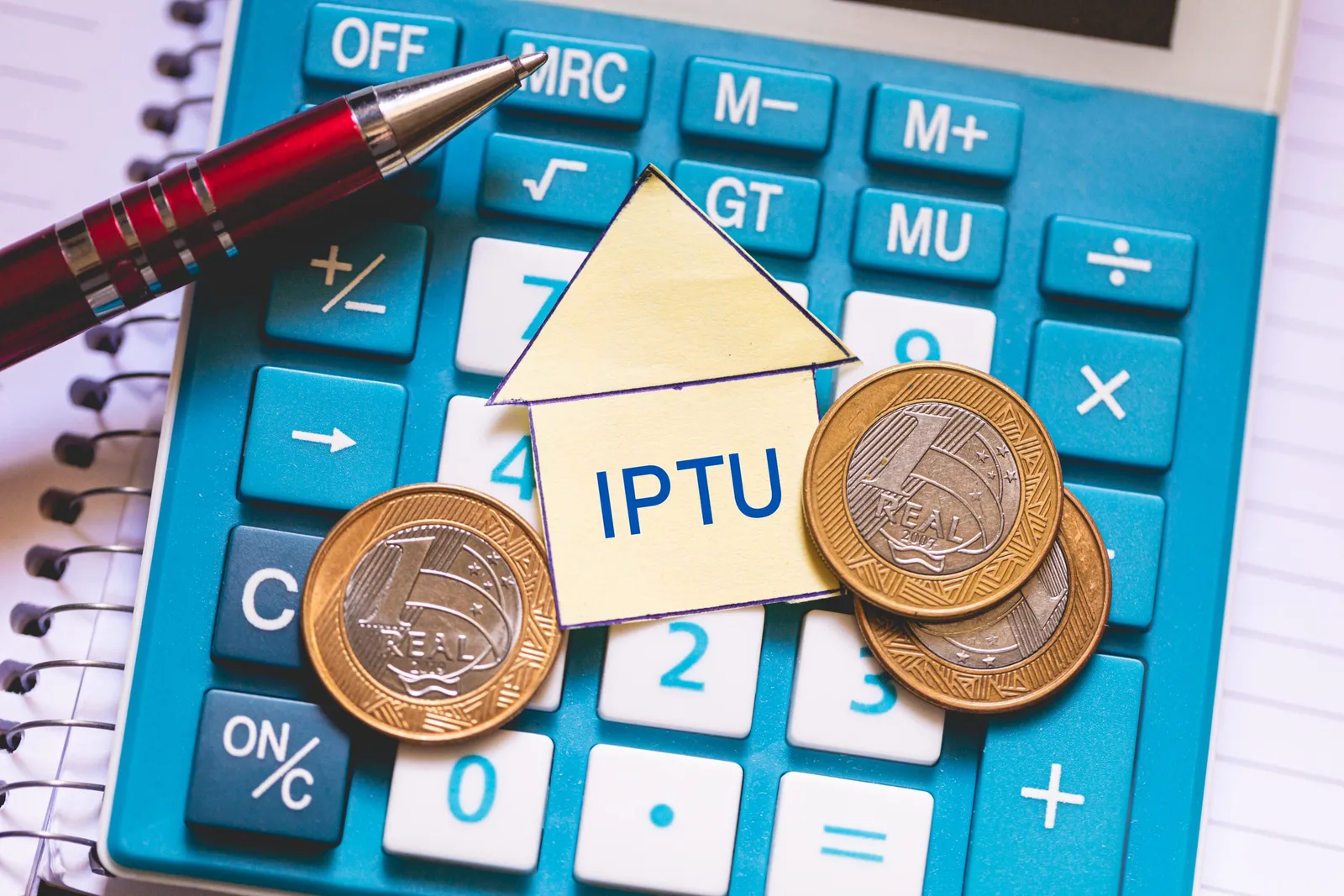 Calculadora azul, caneta, moedas de real e um pequeno papel em formato de casa com a palavra IPTU ilustram o cálculo do imposto após decisão do STF que proíbe cobrança mais cara por tamanho do imóvel.