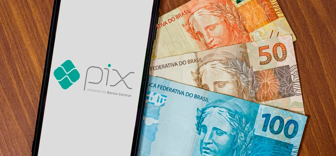 Smartphone exibindo a logo do Pix ao lado de notas de Real brasileiro sobre uma mesa de madeira.