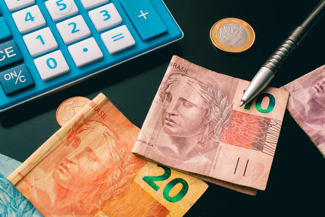 Calculadora, notas de real e caneta sobre uma mesa escura, representando o cálculo de tributos como a exclusão do ISS da base de PIS e COFINS.