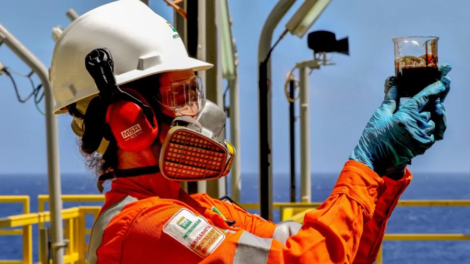 Trabalhador com EPI segura um recipiente com petróleo bruto em uma plataforma de exploração no mar.