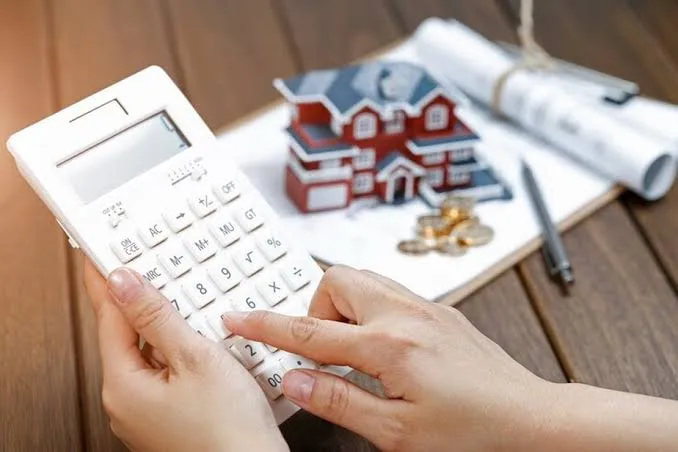 Mãos usando uma calculadora ao lado de uma maquete de casa e moedas, ilustrando o impacto financeiro de que o imposto da herança vai aumentar.