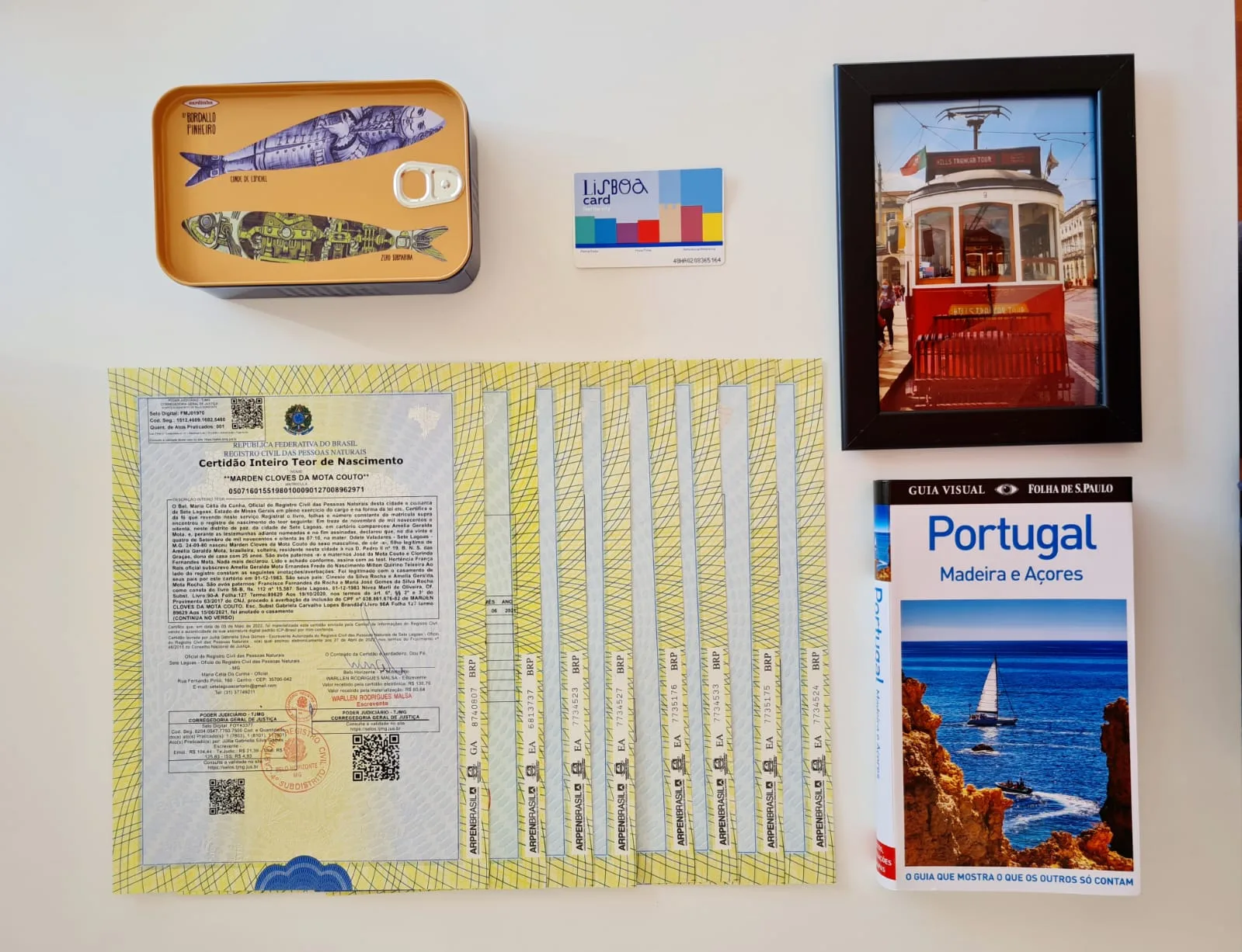 Vista superior de uma mesa com uma Certidão de Inteiro Teor de Nascimento brasileira rodeada por itens de viagem a Portugal, incluindo um guia turístico "Portugal - Madeira e Açores", um porta-retratos com a foto de um bonde elétrico de Lisboa, um cartão "Lisboa Card" e uma lata decorativa.