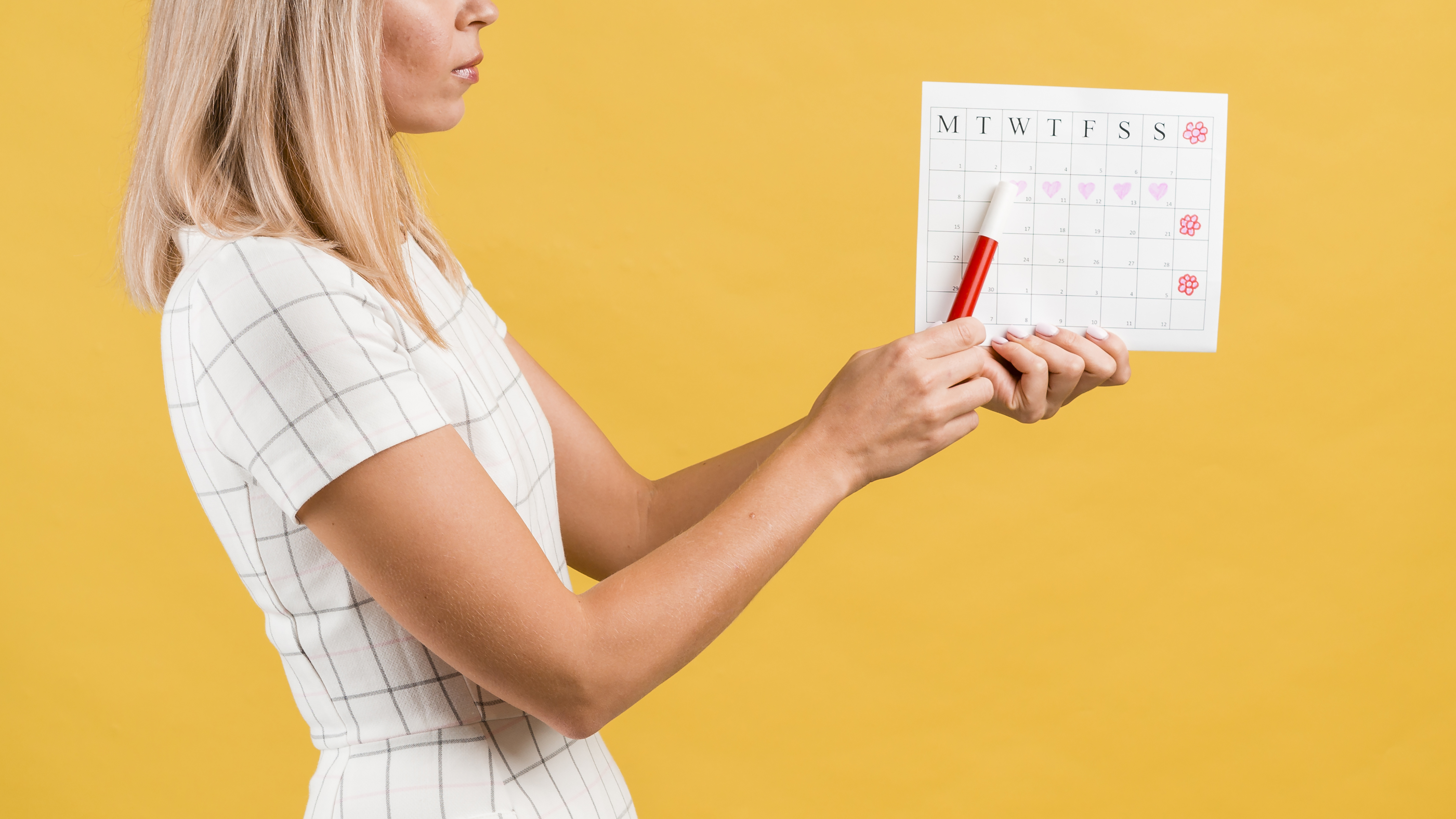 Mulher loira de perfil segura um calendário e uma caneta vermelha contra um fundo amarelo, lembrando prazos e vencimentos que chegam às empresas na última semana de setembro.