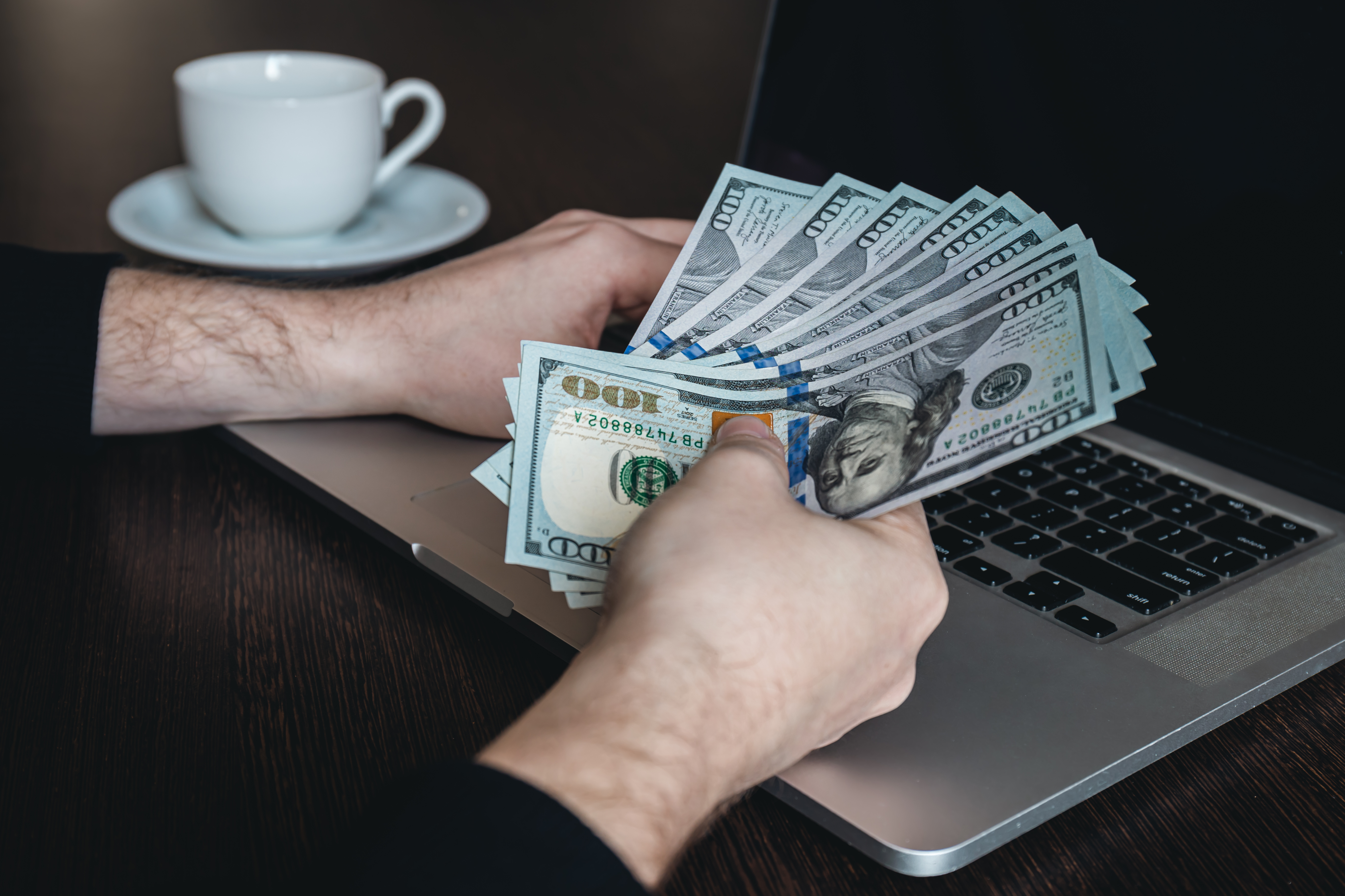 Pessoa segurando notas de dinheiro em leque sobre o teclado de um notebook ao lado de uma xícara de café, ilustrando o controle financeiro para evitar perdas com créditos da CBS na nota fiscal.