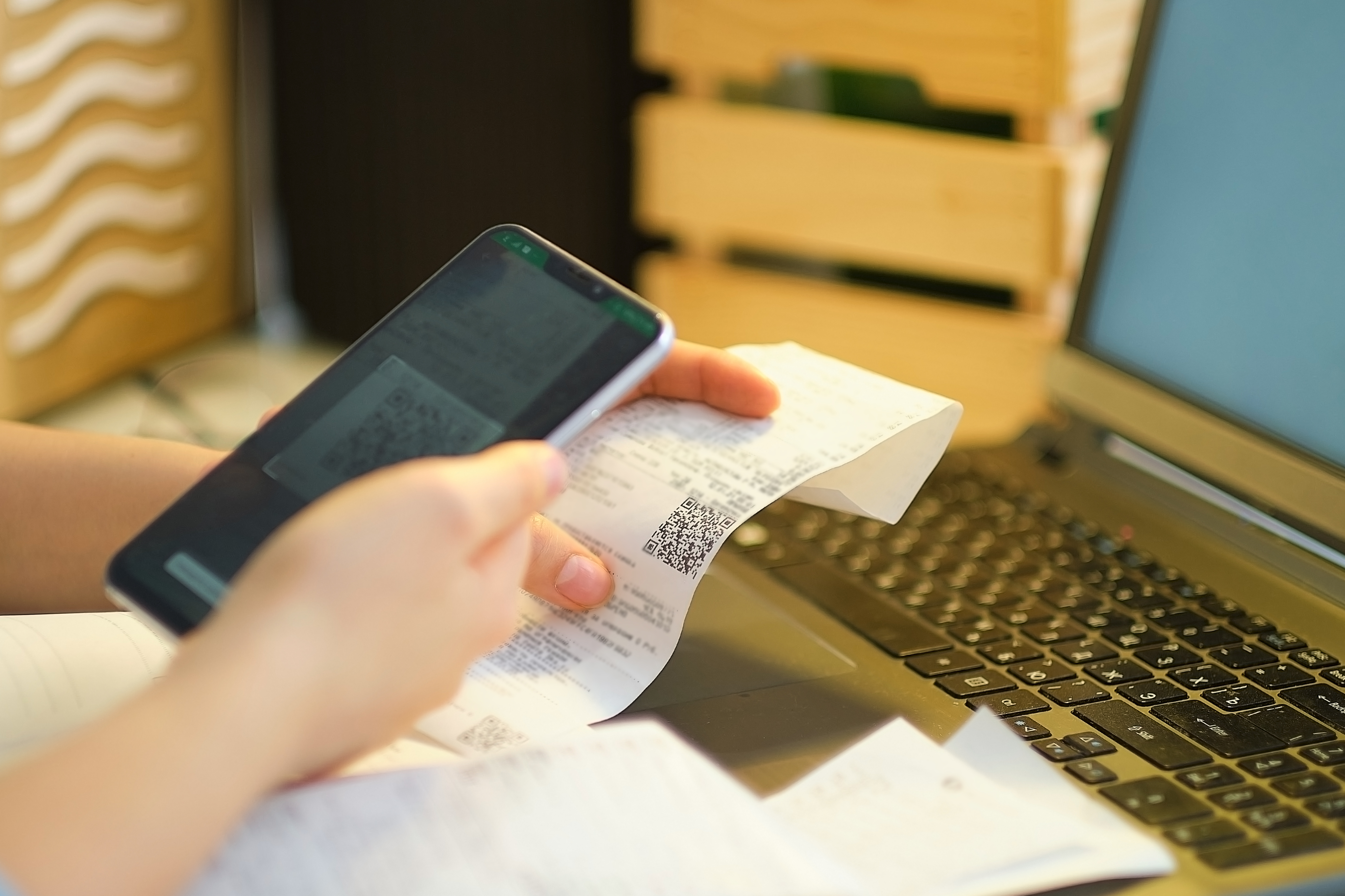 Pessoa usa o celular para escanear o QR code de uma nota fiscal impressa próxima a um notebook, ilustrando a conferência e correção de nota fiscal eletrônica.