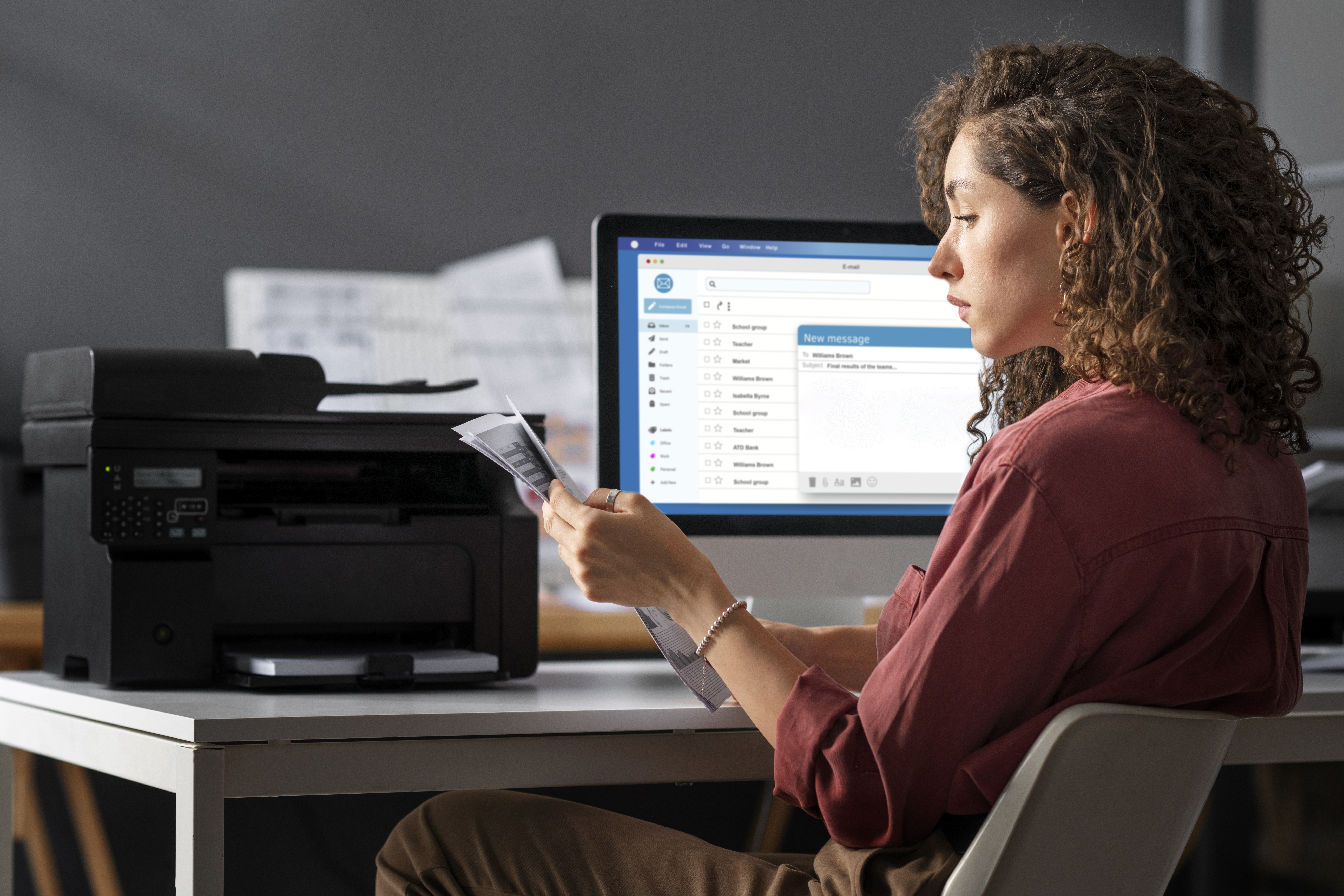 Mulher sentada à escrivaninha analisando documentos impressos e revisando uma nota fiscal eletrônica no computador.