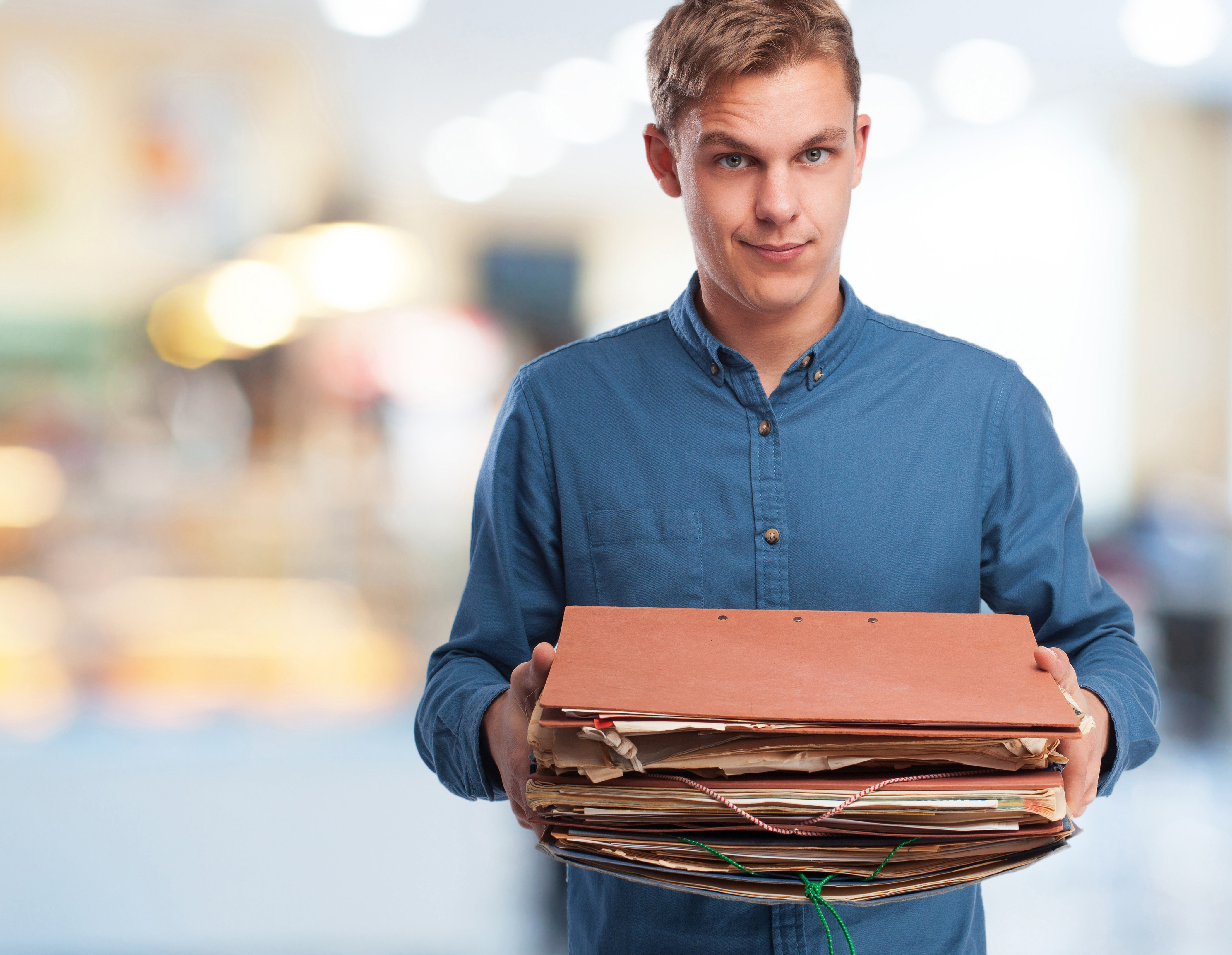 Homem de camisa azul segura uma grande pilha de pastas e documentos, ilustrando a necessidade de guardar notas fiscais por vários anos.