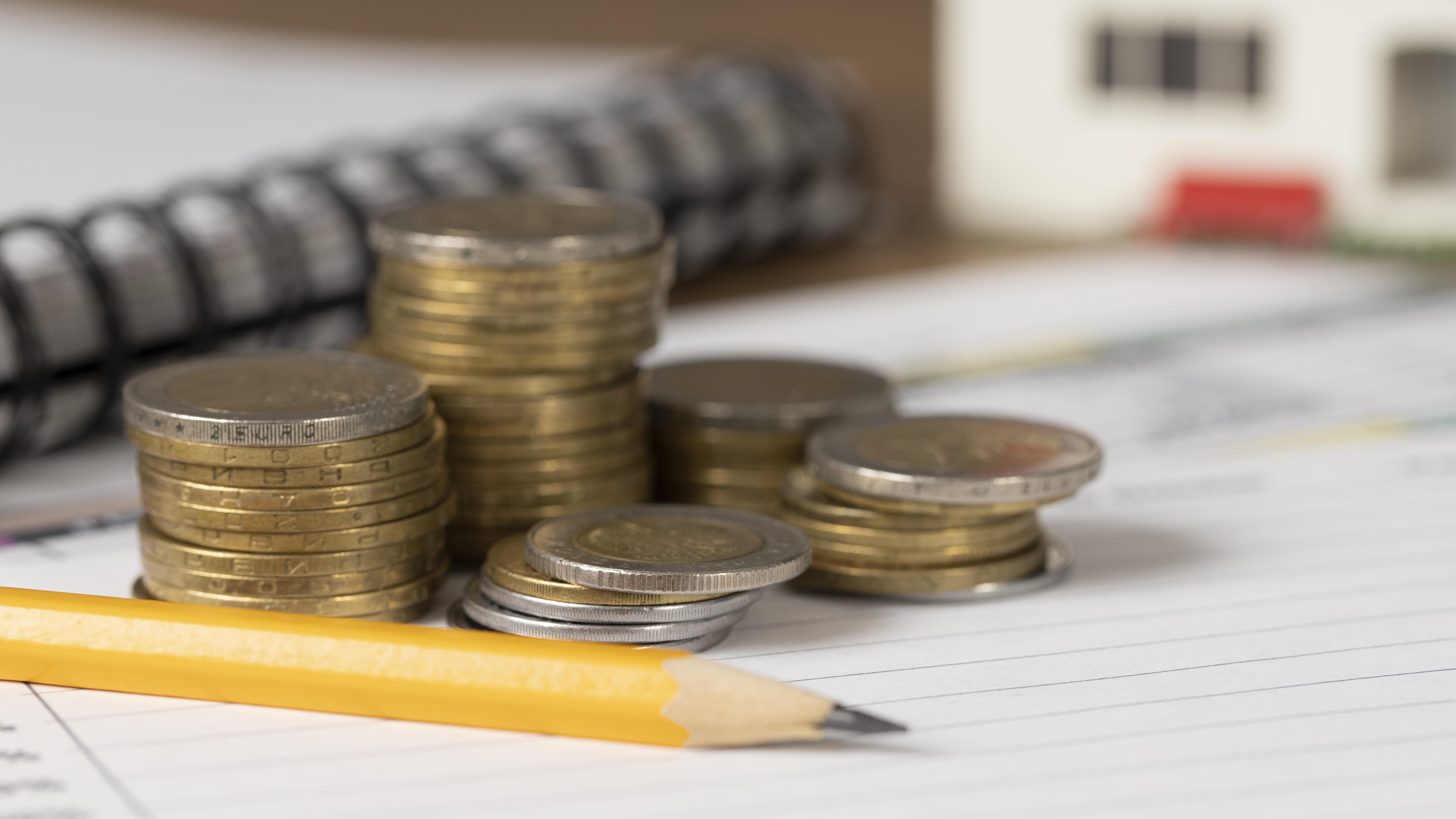 Pilhas de moedas sobre um bloco de notas com um lápis amarelo, simbolizando o impacto financeiro da reforma tributária e do alto IVA.